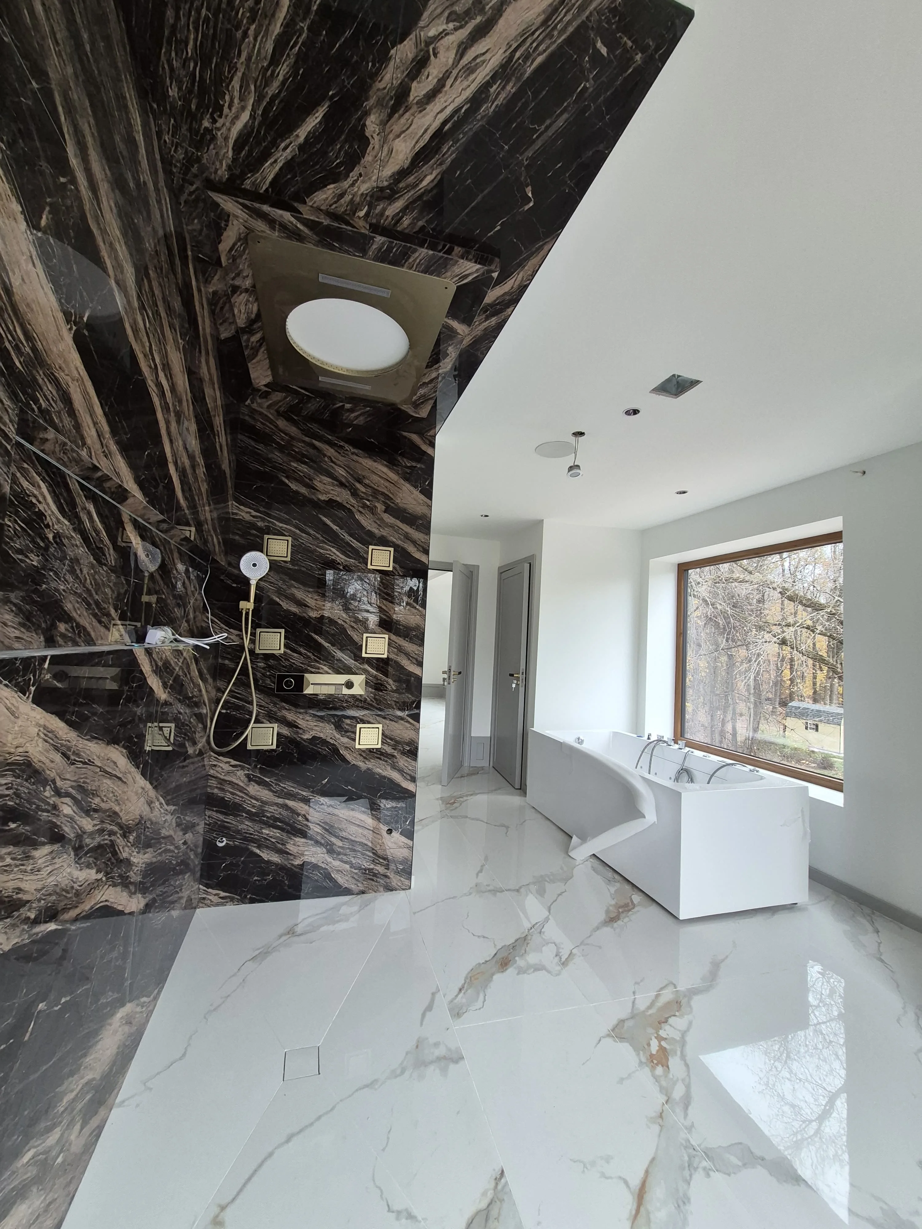 Luxury master bathroom — bookmatched black-and-gold marble shower wall, brass fixtures, freestanding smart-control tub, Calacatta floor